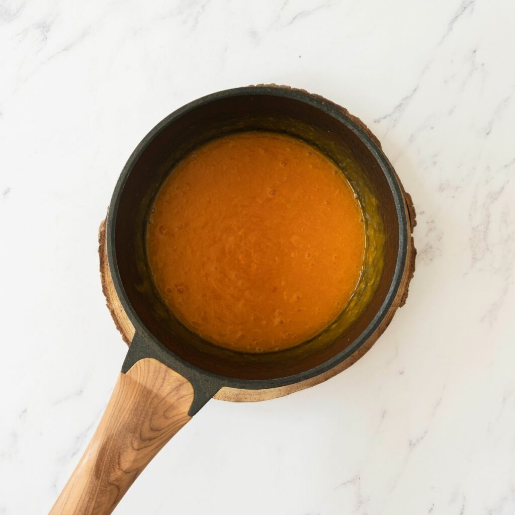 A small saucepan with a wooden handle containing a thick orange sauce sits on a round wooden trivet on a white marble surface.