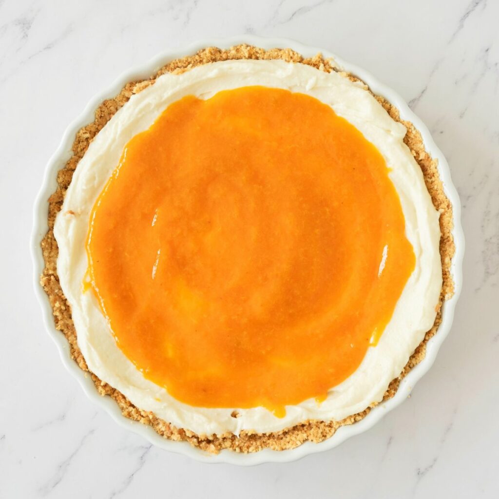 A round pie with a crumb crust, topped with a layer of white cream and an orange glaze, on a white marble surface.