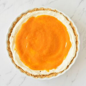 A round pie with a crumb crust, topped with a layer of white cream and an orange glaze, on a white marble surface.