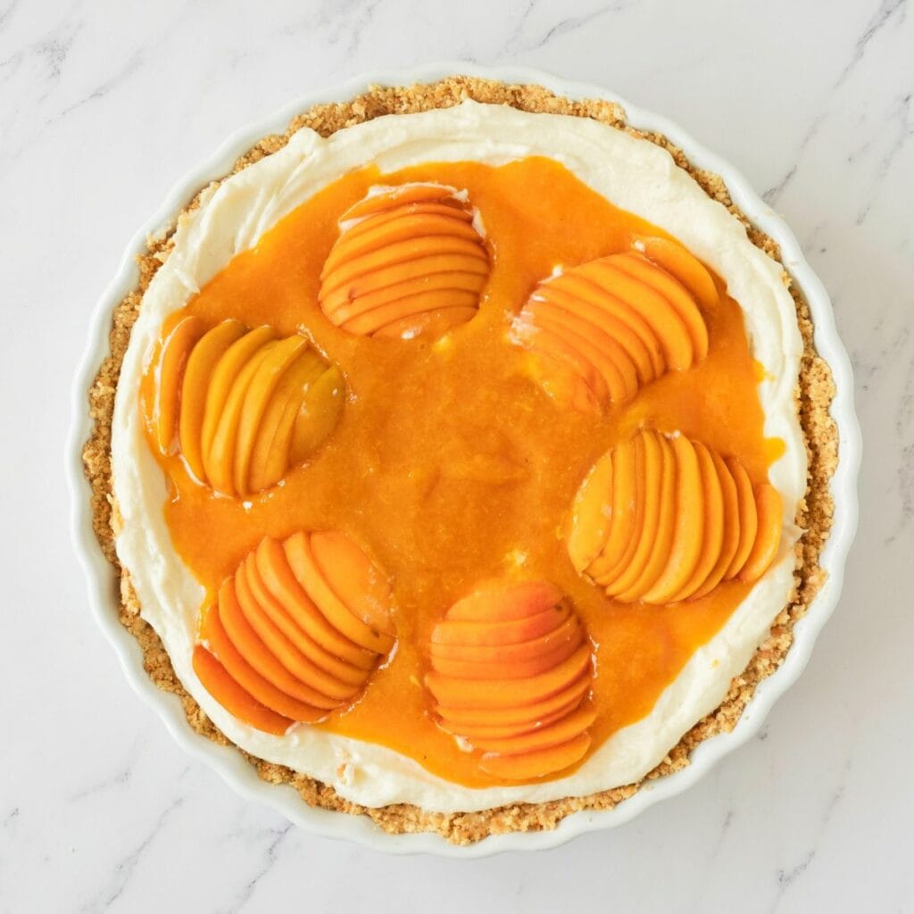 A tart with a crumbly crust, creamy filling, and six neatly sliced peach halves arranged on top with a glossy peach glaze.