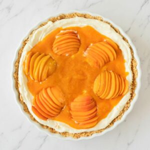 A tart with a crumbly crust, creamy filling, and six neatly sliced peach halves arranged on top with a glossy peach glaze.