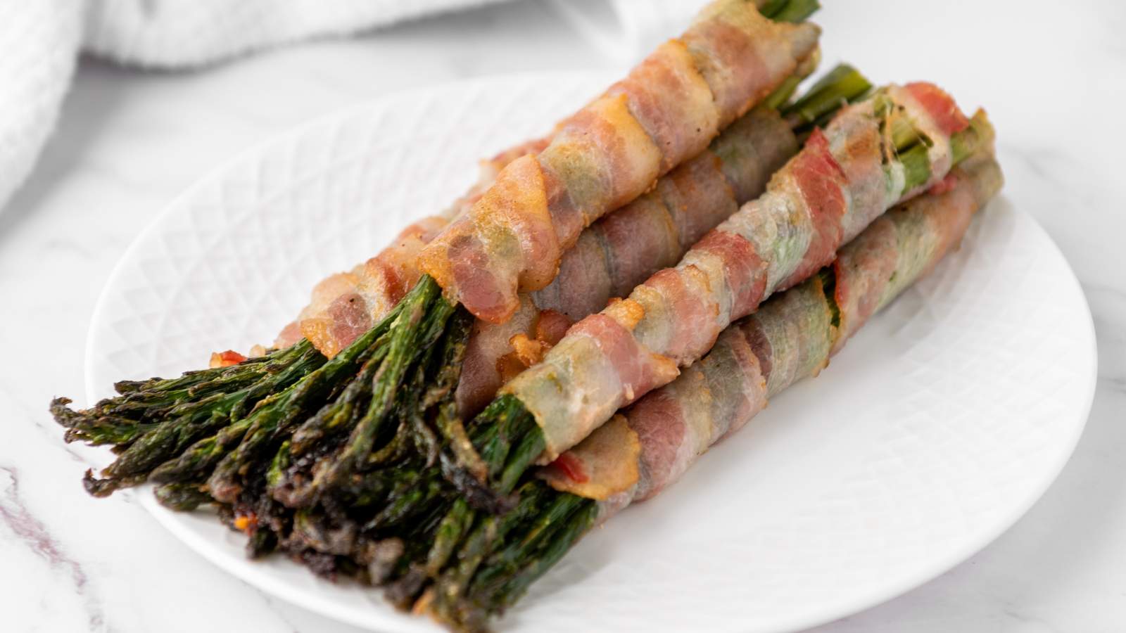 Four bundles of bacon wrapped asparagus are arranged on a white plate.