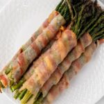 Five bundles of asparagus wrapped in cooked bacon are arranged on a white plate.