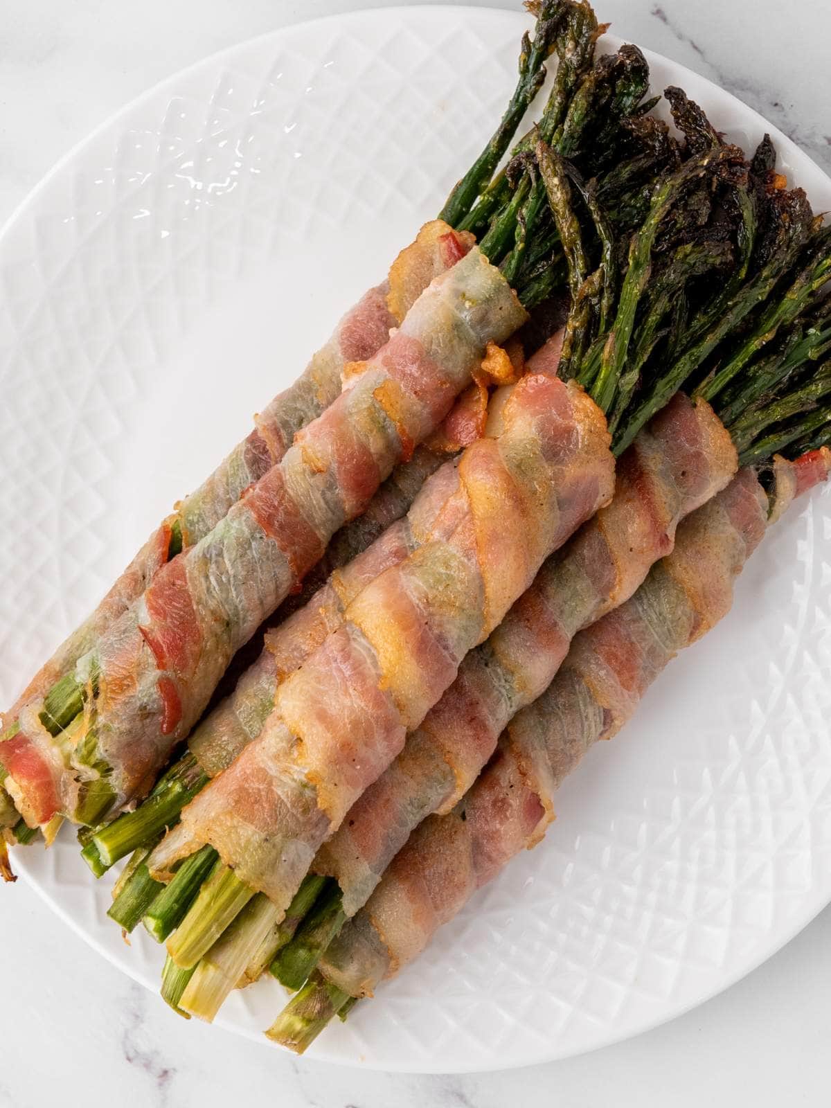 Five bundles of asparagus wrapped in cooked bacon are arranged on a white plate.