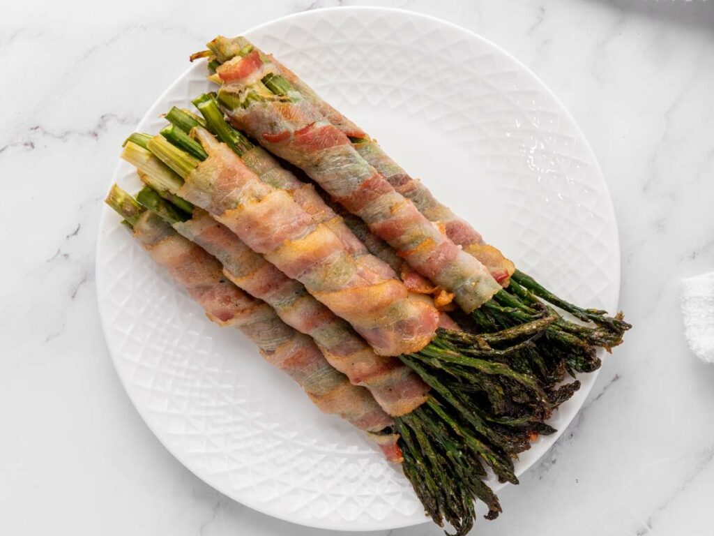 Six bundles of asparagus wrapped in bacon are arranged in a row on a white plate with a textured rim, set on a marble surface.