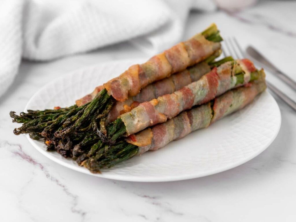 A white plate with several bundles of asparagus spears wrapped in crispy bacon.