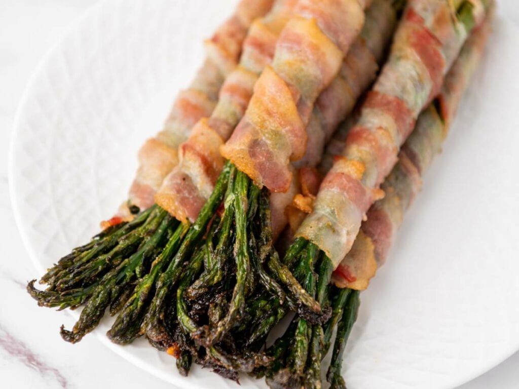 Bacon-wrapped asparagus spears arranged in a row on a white plate.