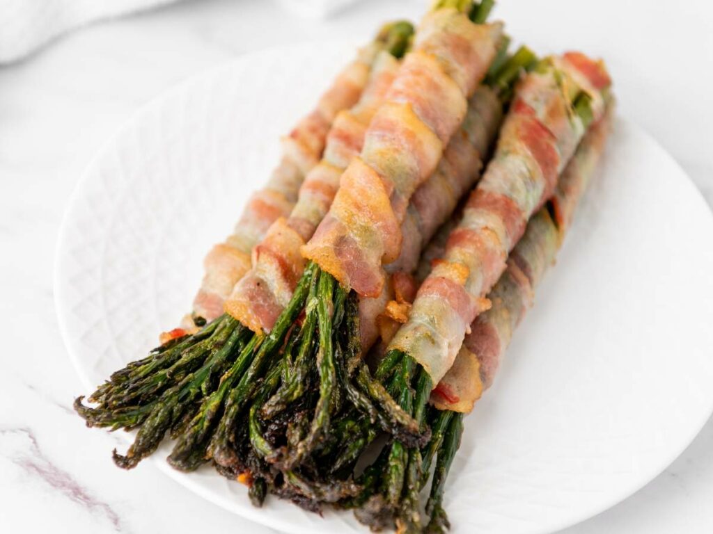 Bundles of asparagus spears wrapped in cooked bacon are arranged on a white plate.