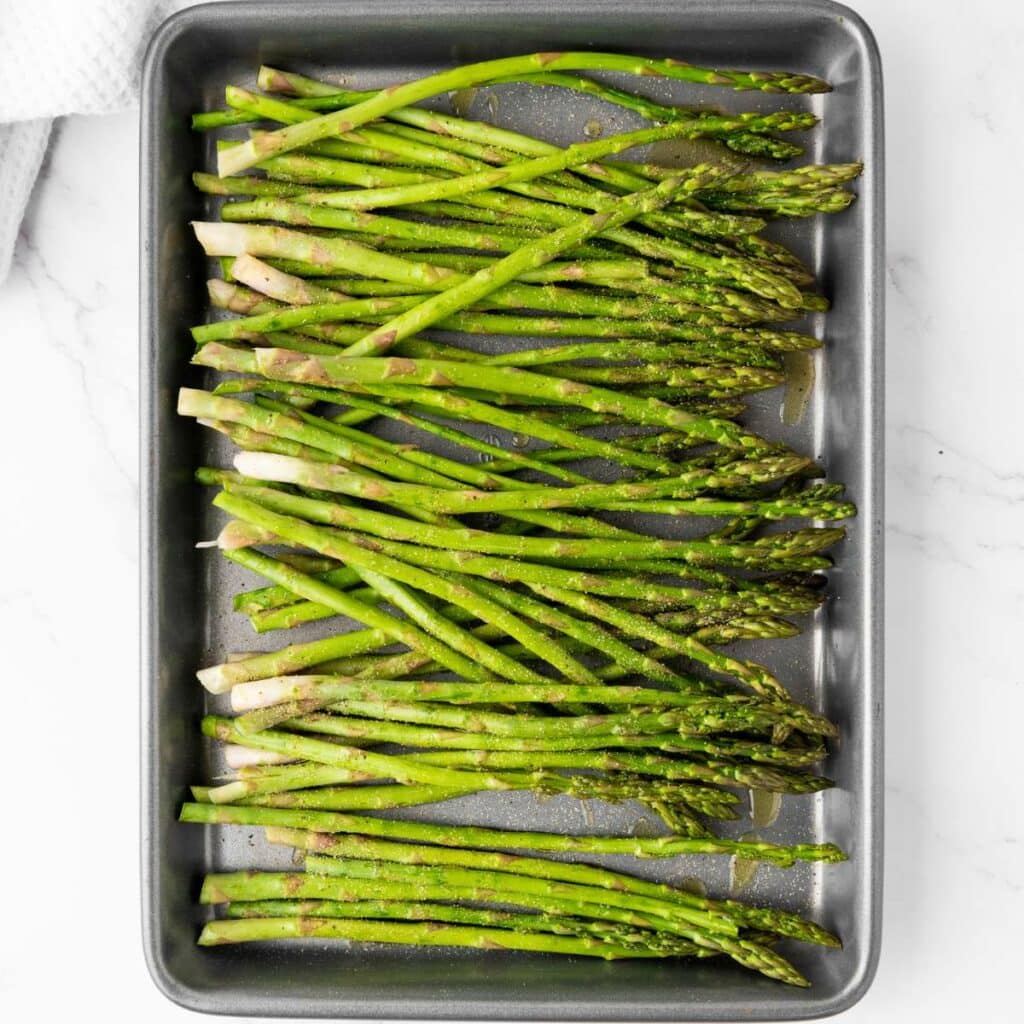 A metal baking tray with raw asparagus spears seasoned with oil and spices, arranged in a single layer on a white surface.