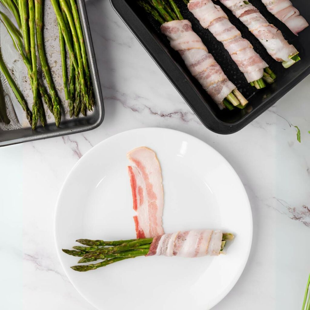 A white plate with raw asparagus spears partially wrapped in bacon, next to a sheet pan of roasted asparagus.