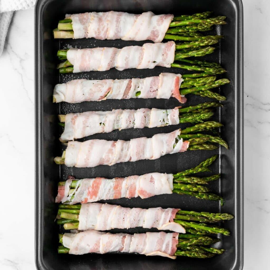 A baking tray with bundles of asparagus spears wrapped in raw bacon, arranged in rows on a white marble surface.