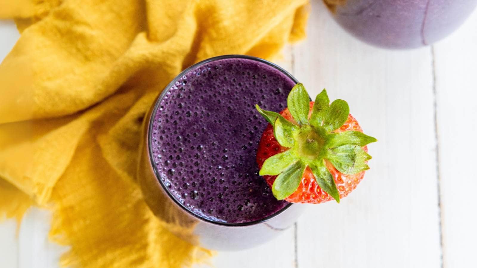 A glass of purple smoothie topped with a whole strawberry, placed on a white surface next to a yellow cloth.