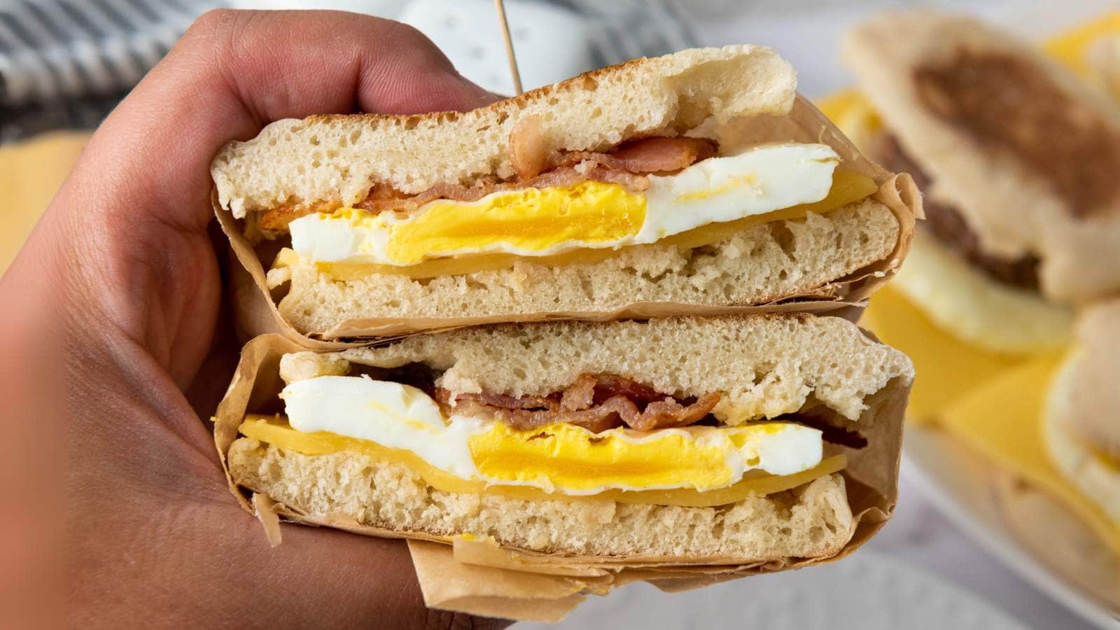 A hand holds a breakfast sandwich cut in half, showing layers of egg, bacon, and cheese between halves of an English muffin.