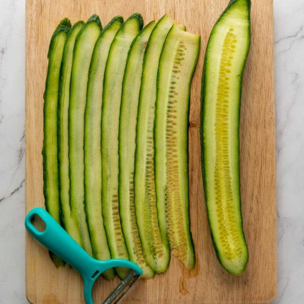 Thin cucumber slices and a blue vegetable peeler are arranged on a wooden cutting board placed on a marble surface.