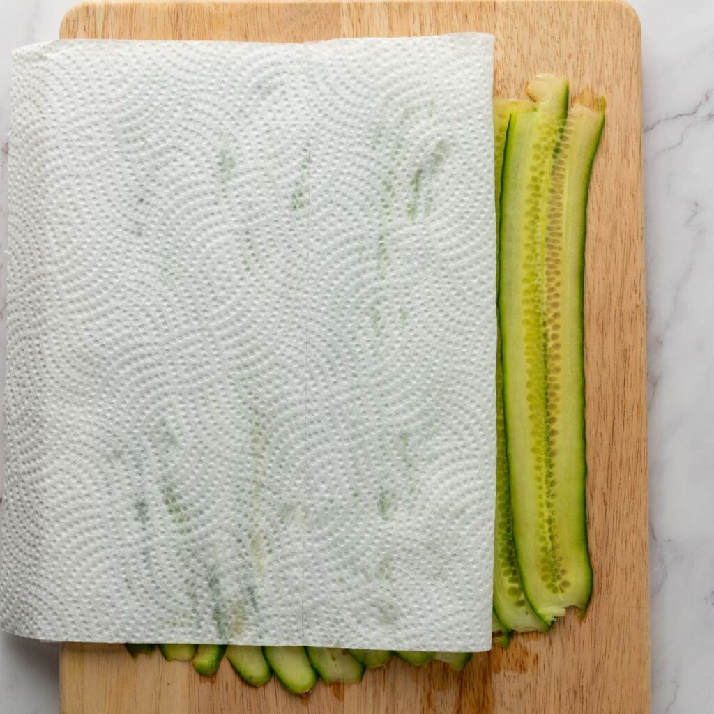 Thin cucumber slices on a wooden cutting board, partially covered by a sheet of patterned paper towel.