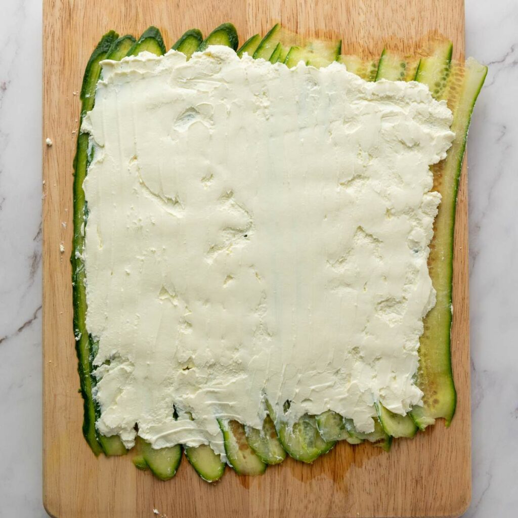Thin cucumber slices are arranged side by side on a wooden board and covered with an even layer of cream cheese.