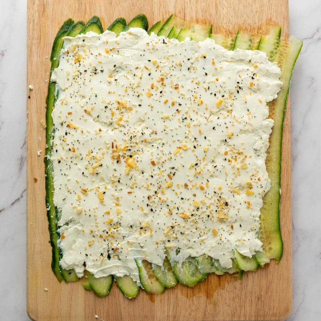 Thin cucumber slices are arranged on a cutting board and topped with a layer of cream cheese sprinkled with seasoning.