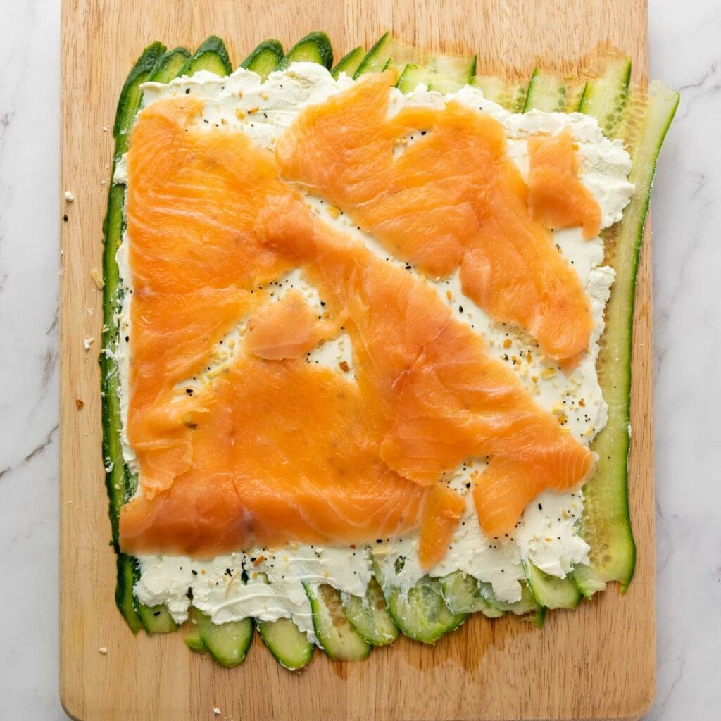Sliced cucumber, cream cheese, and smoked salmon layered on a wooden cutting board.