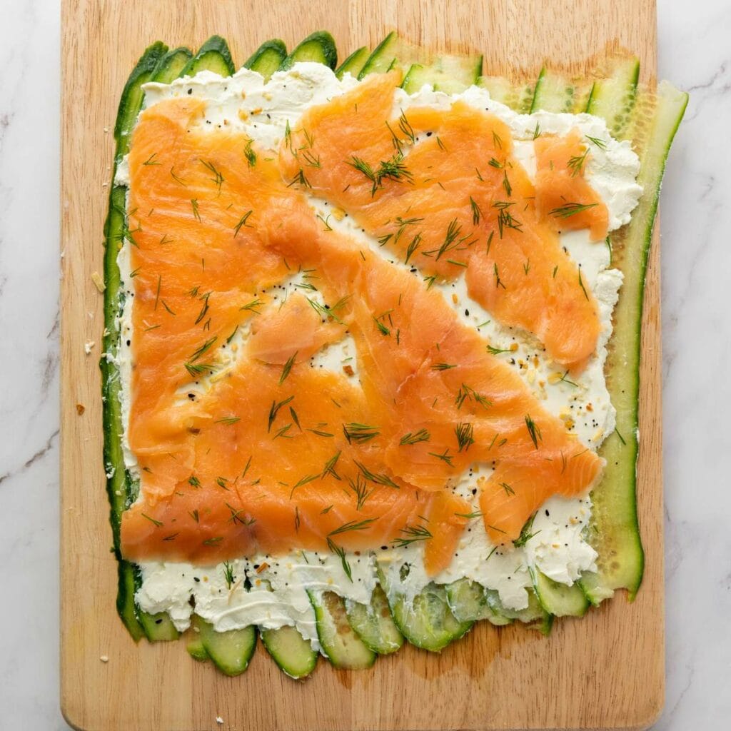 Sliced cucumber layered on a wooden board, topped with a spread of cream cheese.