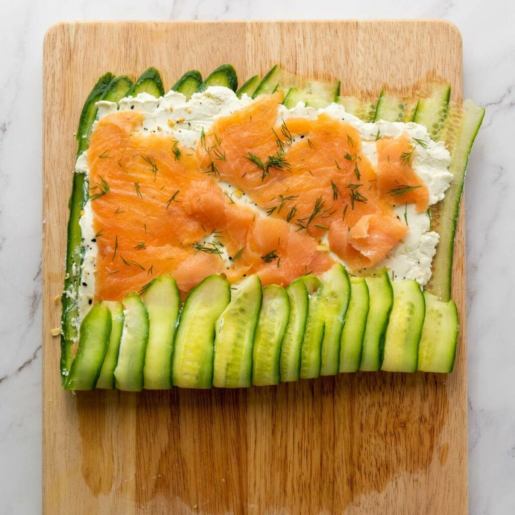 A wooden cutting board with a layer of sliced cucumbers, spread with cream cheese.