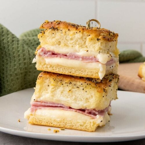 Two stacked ham and cheese sliders on hawaiian rolls on poppy seed bread are displayed on a white plate.