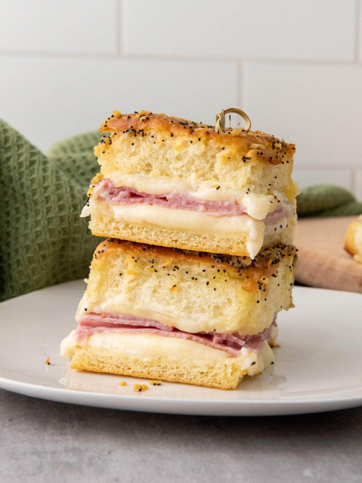 Two stacked ham and cheese sliders on hawaiian rolls on poppy seed bread are displayed on a white plate.