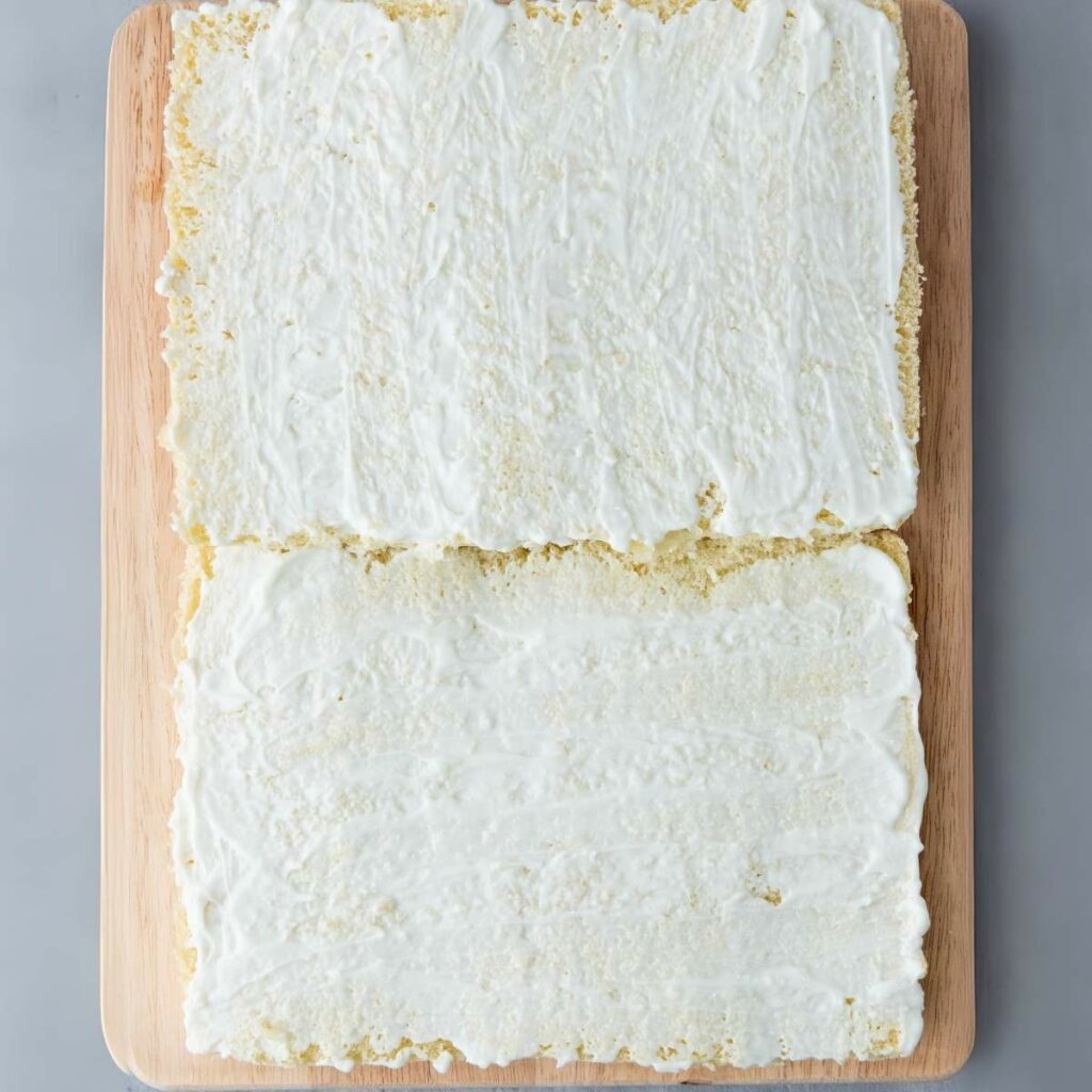 Two rectangular sheets of cake laid side by side on a wooden board, each covered with an even layer of white frosting.