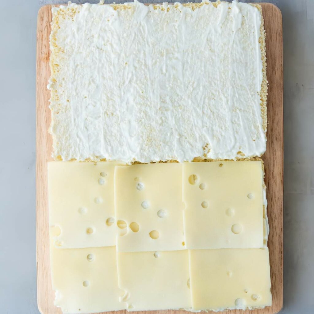 Rectangular sandwich bread on a wooden board, half spread with a white creamy spread and half covered with slices of Swiss cheese.