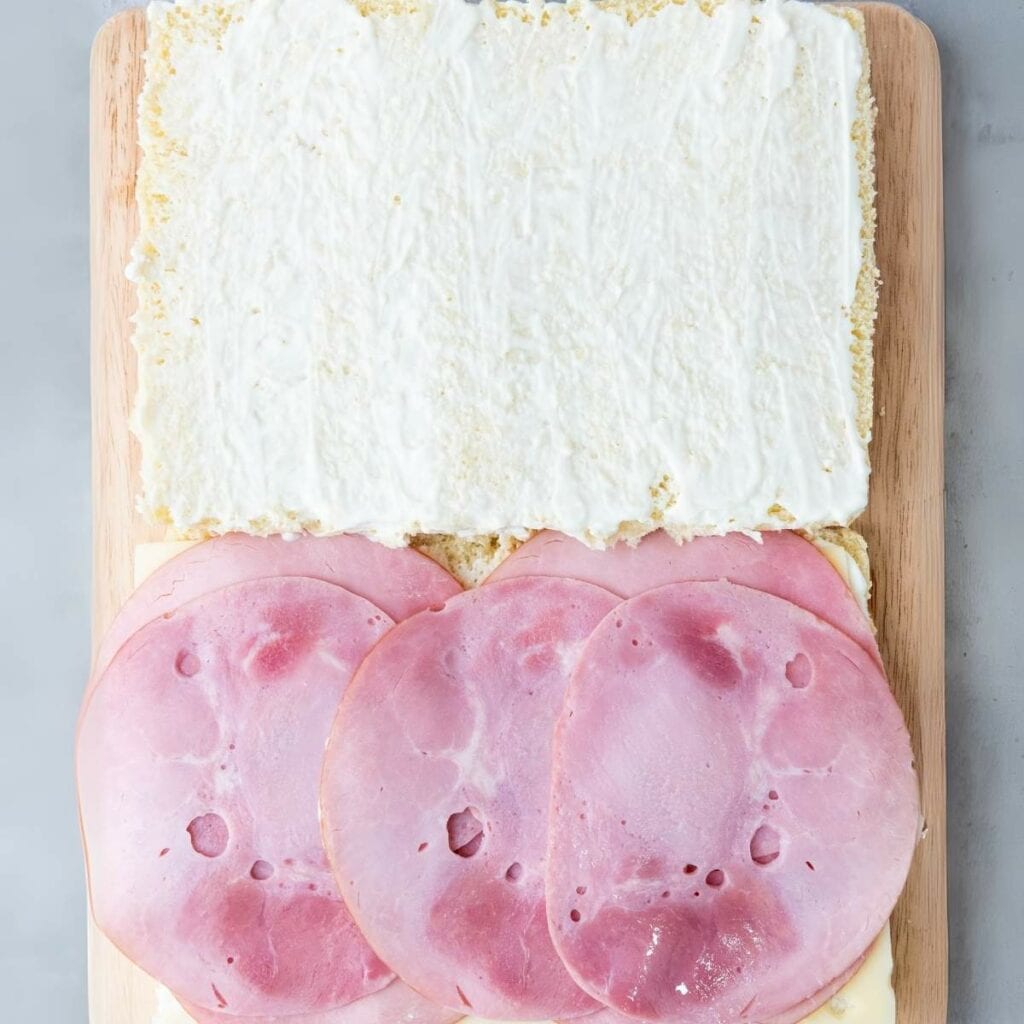 An open sandwich with slices of ham and cheese on bread, topped with a layer of white spread, placed on a wooden board.
