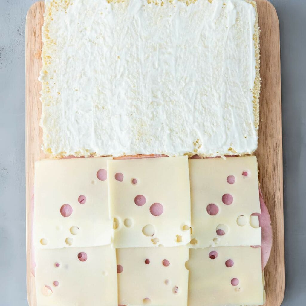 A sandwich being assembled on a wooden board with one half covered in a white spread .