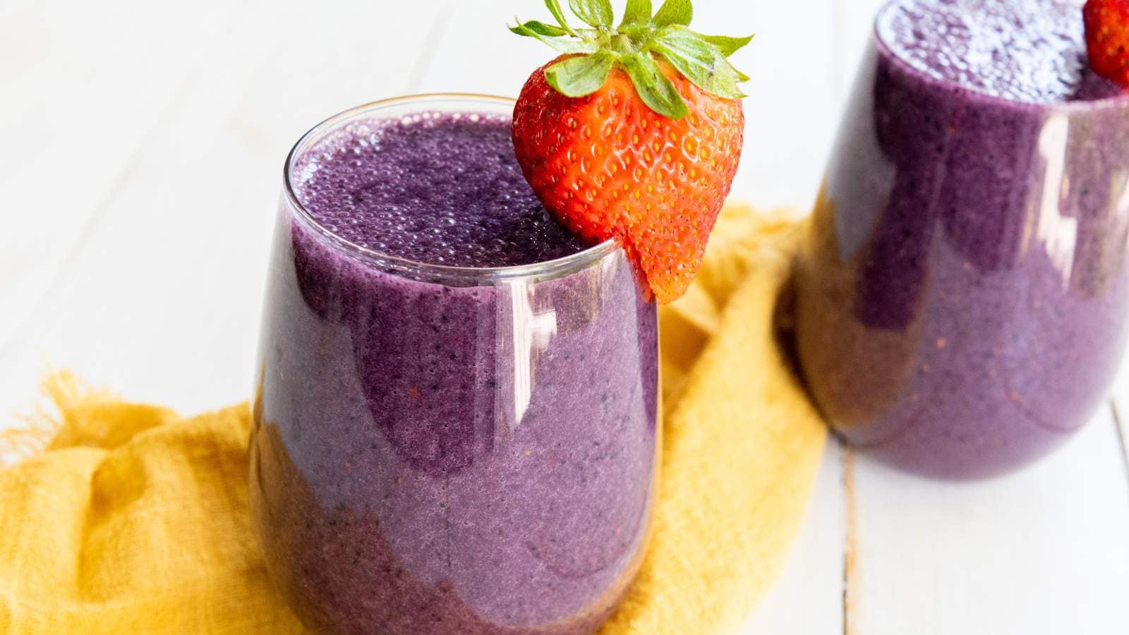 Two glasses of mixed berry smoothie, each garnished with a whole strawberry, are placed on a yellow cloth on a light wooden surface.