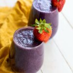 Two glasses of mixed berry smoothie garnished with whole strawberries, placed on a yellow cloth on a white surface.