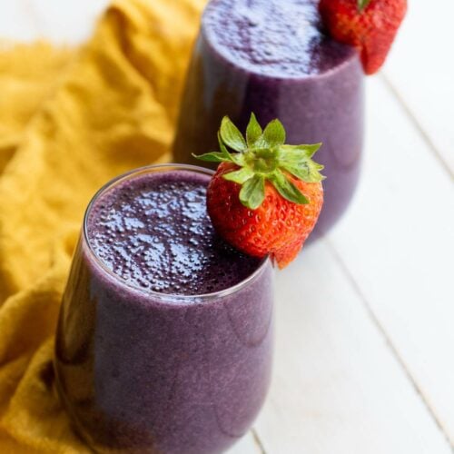 Two glasses of mixed berry smoothie garnished with whole strawberries, placed on a yellow cloth on a white surface.