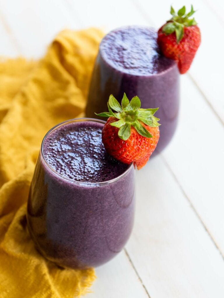 Two glasses of mixed berry smoothie garnished with whole strawberries, placed on a yellow cloth on a white surface.