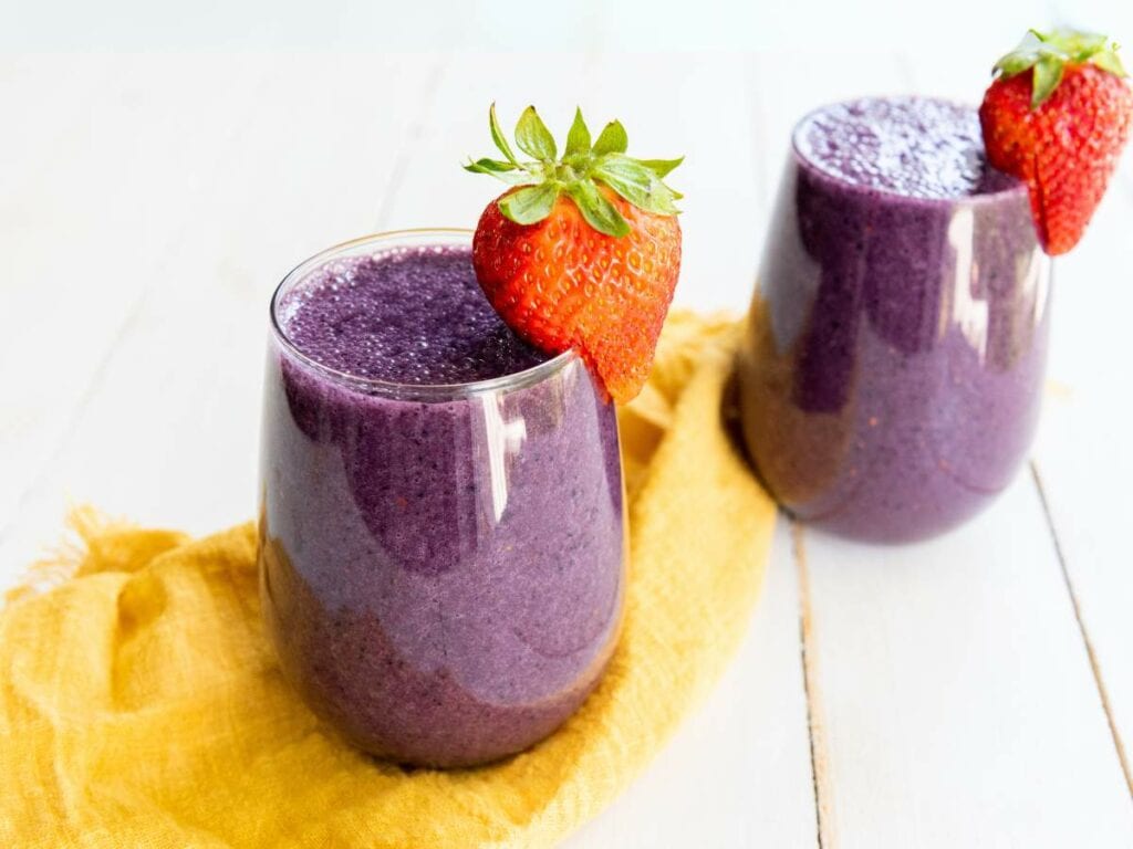 Two glasses of mixed berry smoothie garnished with fresh strawberries, placed on a white surface with a yellow cloth underneath.