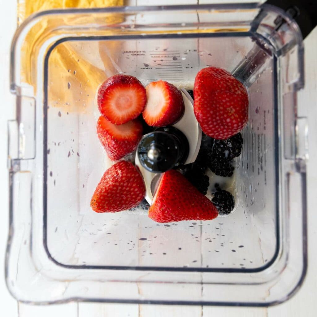 Top-down view of a blender containing several strawberries and blackberries, ready to be blended.