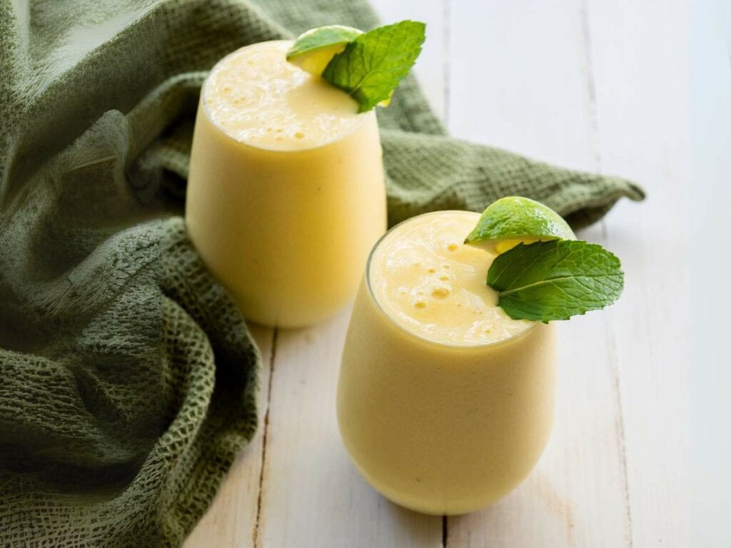 Two glasses of pineapple and banana smoothie topped with mint leaves sit on a white wooden surface.