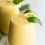 A glass of pineapple and banana smoothie garnished with a lime wedge and mint leaf.