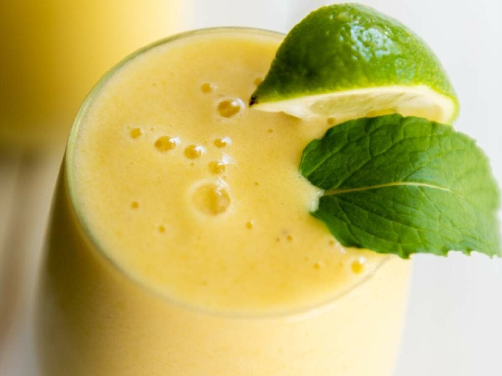 A close-up of a pineapple and banana smoothie in a glass, garnished with a lime wedge and fresh mint leaf.