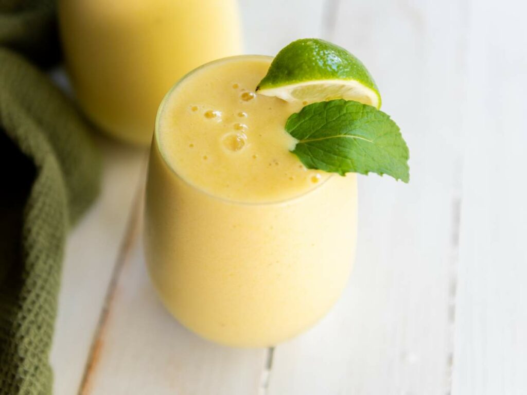 A glass of pineapple and banana smoothie garnished with a lime wedge and mint leaf sits on a white wooden surface.