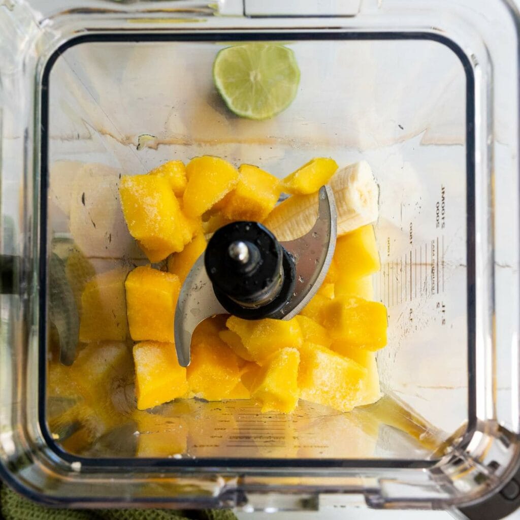 Chunks of frozen mango and a piece of banana in a blender, with half a lime resting at the top edge of the blender.
