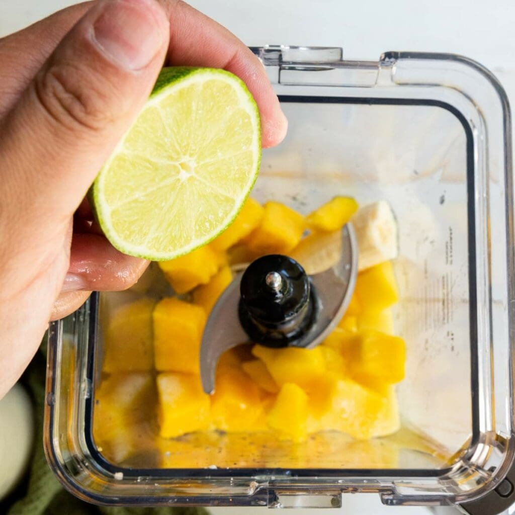 A hand holds half a lime over a blender containing chopped mango and banana pieces, ready to be blended.