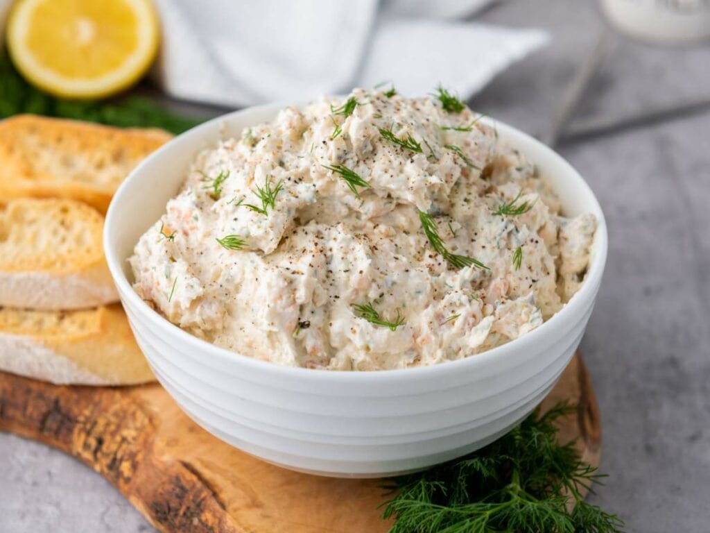 A white bowl filled with smoked salmon spread garnished with fresh dill, set on a wooden board.