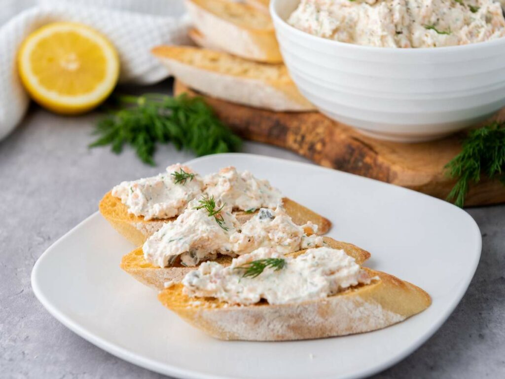 Three slices of toasted baguette topped with smoked salmon spread and dill on a white plate.