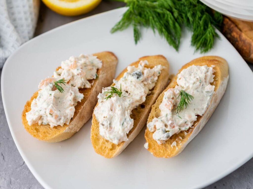 Three pieces of toasted bread topped with a creamy spread and garnished with dill, served on a white plate.