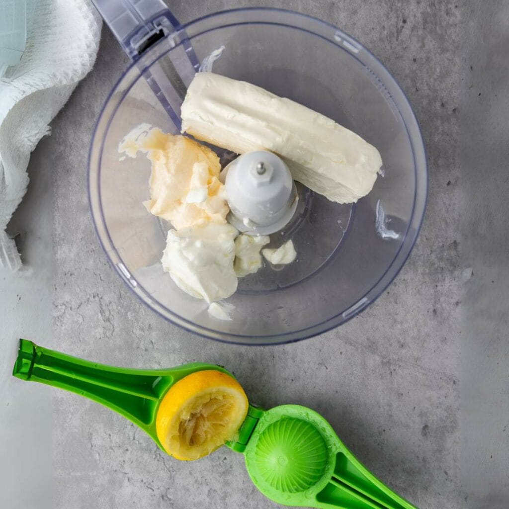 A food processor bowl with cream cheese and mayonnaise inside, next to a green lemon squeezer holding a halved lemon on a gray countertop.