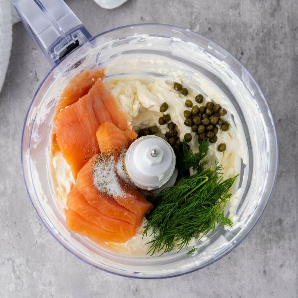 A food processor bowl containing smoked salmon, cream cheese, capers, fresh dill, and salt, ready for blending, on a gray countertop.