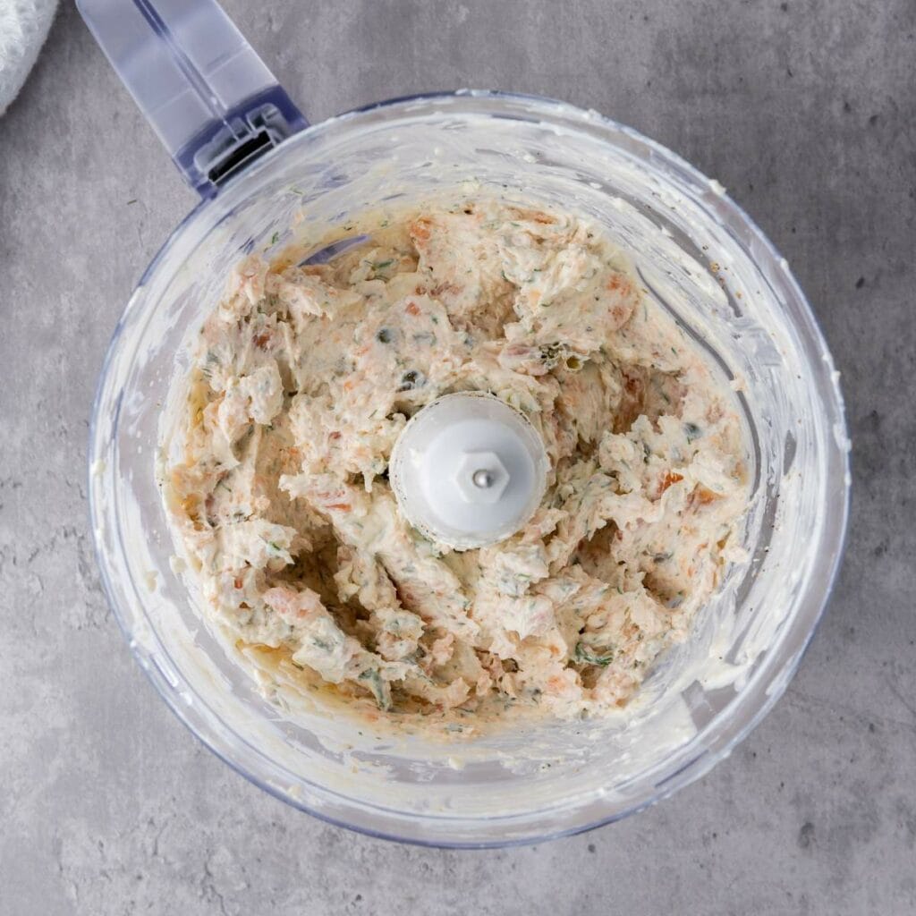 A food processor bowl containing a creamy, mixed spread with visible herb and spice flecks sits on a gray countertop.