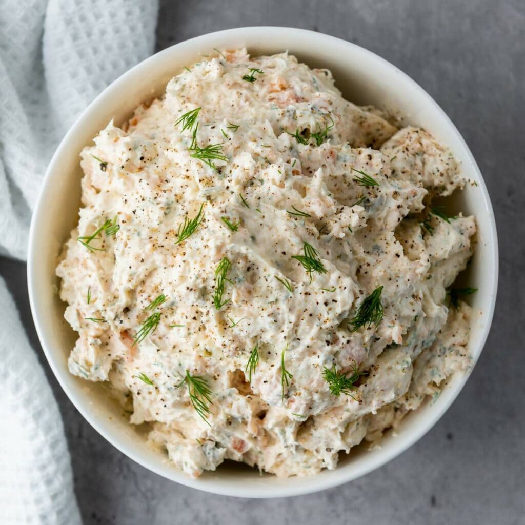 A bowl of creamy dip topped with fresh dill and black pepper, placed on a gray surface next to a textured white cloth.