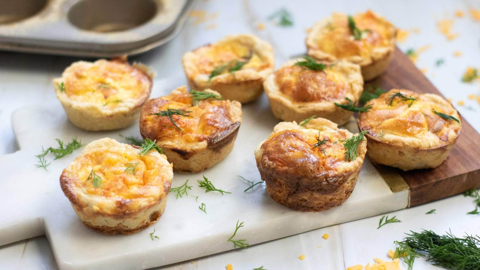 Smoked salmon tartlets with golden tops are arranged on a white cutting board.